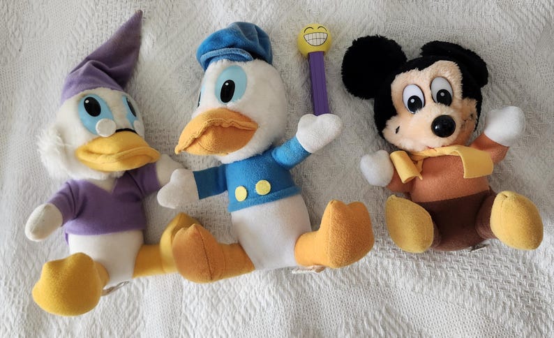 Puede incluir: Tres peluches de personajes de Disney: Pato Donald, Mickey Mouse y una variante de Pato Donald. Pato Donald viste camisa y gorro de marinero azules, Mickey Mouse tiene cuerpo marr&oacute;n y orejas negras. La variante de Pato Donald viste un atuendo morado.