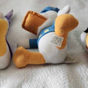 Puede incluir: Tres peluches de personajes de Disney: Pato Donald, Pata Daisy y Goofy. El Pato Donald lleva una camisa y un gorro de marinero azules. Pata Daisy lleva un top morado. Goofy lleva un atuendo marr&oacute;n. Todos est&aacute;n tumbados sobre una superficie blanca.