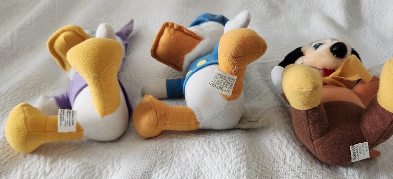 Puede incluir: Tres peluches de personajes de Disney: Pato Donald, Pata Daisy y Goofy. El Pato Donald lleva una camisa y un gorro de marinero azules. Pata Daisy lleva un top morado. Goofy lleva un atuendo marr&oacute;n. Todos est&aacute;n tumbados sobre una superficie blanca.