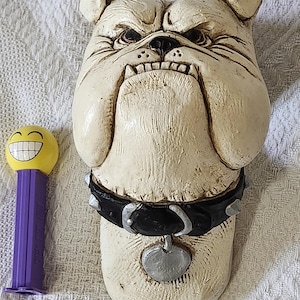 Può includere: Una scultura di bulldog color crema con un collare nero borchiato e un'etichetta a forma di cuore. Un dispenser PEZ giallo e viola con una faccia sorridente è accanto alla scultura.