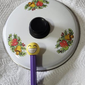 Op de afbeelding: Een witte pannendeksel met een zwarte handgreep en een patroon van kleurrijke groenten. Een gele PEZ-dispenser met een smiley-gezicht en een paarse basis staat voor het deksel.