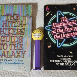 May include: Two paperback books, "The Hitch-Hiker's Guide to the Galaxy" and "The Restaurant at the End of the Universe", with a yellow and purple Pez dispenser. The first book has a colorful cover, the second a black cover with neon lights.