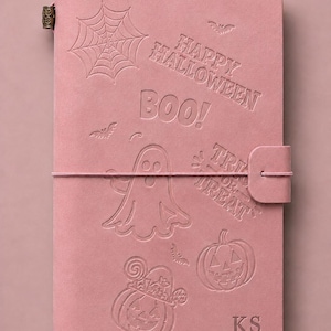 May include: A dusty pink notebook featuring embossed Halloween designs including a spider web, a ghost, two jack-o'-lanterns, bats, and the text "HAPPY HALLOWEEN", "BOO!", and "TRICK OR TREAT". The cover includes a metal clasp and the initials "KS".