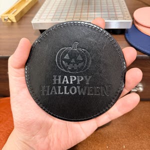 Pode incluir: Um porta-copos circular de couro preto com um gráfico de abóbora de Halloween em relevo e o texto "HAPPY HALLOWEEN" centralizado na superfície. O porta-copos tem costuras visíveis ao longo da borda externa e um acabamento fosco.