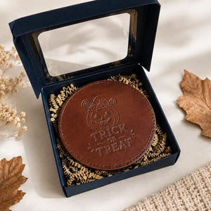 May include: A round brown leather coaster featuring an embossed jack-o'-lantern design with a lollipop and the text "TRICK OR TREAT" inside a dark blue gift box with a clear window lid, nestled on a bed of shredded brown paper filler.