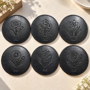 Puede incluir: Un juego de seis posavasos circulares negros de material texturizado, cada uno con un diseño floral en relieve único y las letras BZ en la parte inferior. Las flores representadas incluyen un clavel, un racimo de flores pequeñas, un narciso, una margarita, lirio de los valles y una rosa, todos con bordes cosidos.