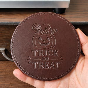 May include: A round, dark brown leather coaster featuring a debossed design of a jack-o'-lantern filled with sweets and the text TRICK OR TREAT. The coaster has visible stitching along the circular edge and a smooth, matte finish on the surface.