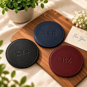 May include: Three circular coasters in black, navy blue, and burgundy, each featuring stitched edges and embossed lettering. The black coaster displays KYS, the navy blue coaster shows A&B, and the burgundy coaster features BZ. A small card reads For You.