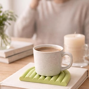 Op de afbeelding: Een gespikkelde witte keramische mok gevuld met een warme drank, rustend op een lichtgroene, geribbelde siliconen onderzetter. De onderzetter staat op een gesloten boek. Een brandende kaars en een persoon die een mok vasthoudt, zijn op de achtergrond.