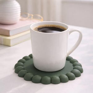 Op de afbeelding: Een witte keramische mok gevuld met donkere koffie staat op een donkergroene onderzetter met geschulpte rand. De mok heeft een gebogen oor. De onderzetter staat op een wit oppervlak. Een vaas met witte bloemen en boeken staan op de achtergrond.