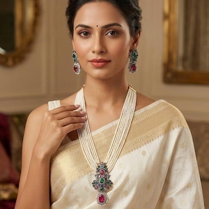 Op de afbeelding: Een meerstrengs witte parelketting met een grote, versierde hanger met rode en groene edelstenen. Bijpassende oorbellen met rode en groene stenen vullen de ketting aan. De sieraden worden gedragen bij een crèmekleurig kledingstuk.