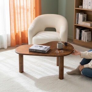May include: A kidney-shaped coffee table made of dark brown wood with three thick cylindrical legs. On the tabletop sits a book titled A Tree in the House, a light grey ceramic mug, and a pair of gold-rimmed glasses with clear lenses.