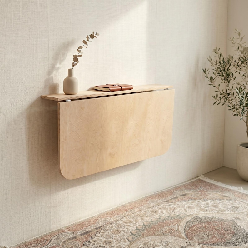 May include: A light-colored wooden wall-mounted drop-leaf table with rounded bottom corners. A small beige ceramic vase containing a single dried branch and a closed brown leather-bound notebook are placed on the flat top surface of the table.