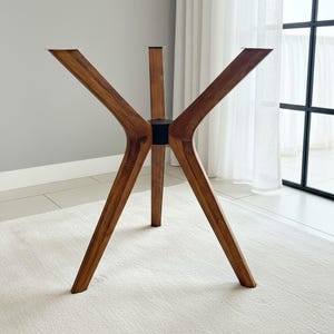May include: A table base featuring four angled wooden legs with a visible grain pattern, connected by a central black cylindrical hub. Each leg terminates in a flat metal mounting plate at the top, designed to support a tabletop surface.