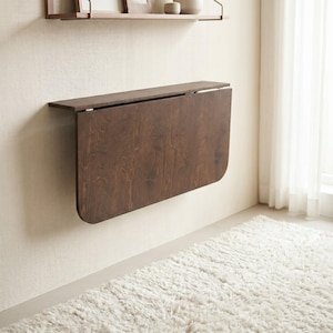 May include: A wall-mounted, dark brown wood-grain drop-leaf table in a folded, vertical position. The rectangular table features rounded corners on the bottom edge and is attached to the wall with a horizontal hinge mechanism.