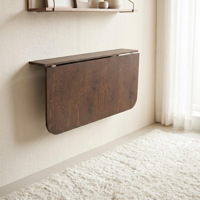 May include: A wall-mounted, dark brown wood-grain drop-leaf table in a folded, vertical position. The rectangular table features rounded corners on the bottom edge and is attached to the wall with a horizontal hinge mechanism.