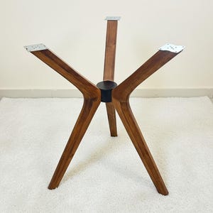 May include: A four-legged table base constructed from medium-toned wood with a visible grain pattern. The legs extend outward from a central black cylindrical hub, each topped with a flat, square silver metal mounting plate for attachment.