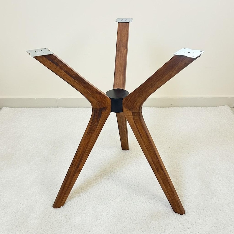 May include: A four-legged table base constructed from medium-toned wood with a visible grain pattern. The legs extend outward from a central black cylindrical hub, each topped with a flat, square silver metal mounting plate for attachment.