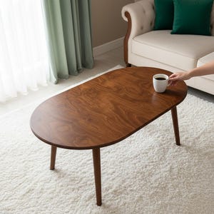 Op de afbeelding: Ovale salontafel van hout met een donkerbruine afwerking. De tafel heeft vier taps toelopende poten en een glad oppervlak. Een witte mok met koffie staat op de tafel. De tafel staat op een wit hoogpolig tapijt.