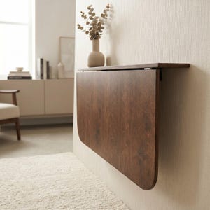 May include: A wall-mounted, dark brown wood-grain drop-leaf table featuring a rectangular top and a rounded-edge folding panel. The table is attached to a light-colored wall with metal hinges, allowing the surface to fold down vertically.