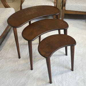 Puede incluir: Juego de tres mesas nido con acabado marrón oscuro. Cada mesa tiene una tapa en forma de media luna y patas cónicas. La mesa más grande mide aproximadamente 76 cm de largo.