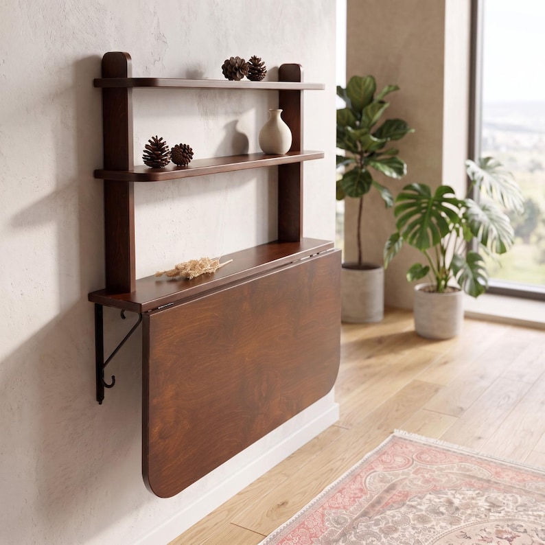 May include: A dark brown wooden wall-mounted unit featuring two upper shelves and a drop-leaf table surface. The top shelf holds two pinecones, while the middle shelf displays two pinecones and a small cream-colored vase. The table is supported by metal brackets.