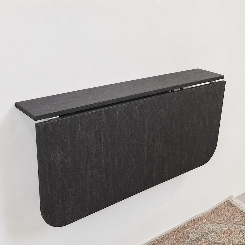 May include: A wall-mounted, charcoal-colored wooden drop-leaf table featuring a rectangular top shelf and a large, vertically oriented panel with rounded bottom corners. The surface displays a visible dark wood grain texture throughout the piece.