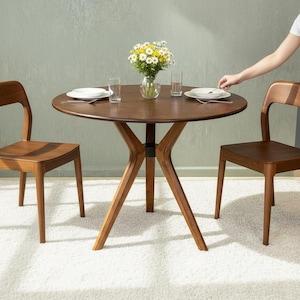 May include: A round dark wood dining table with a central tripod base and two matching wooden chairs. On the table are two white plates, two clear glass tumblers, and a glass vase filled with a bouquet of yellow and white daisies.