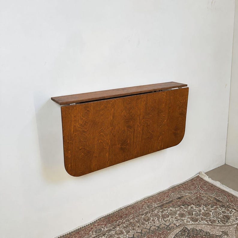 May include: A wall-mounted drop-leaf table featuring a medium-brown wood grain finish. The rectangular top is attached to a hinged, semi-circular drop leaf that hangs vertically against the wall when in the folded position.