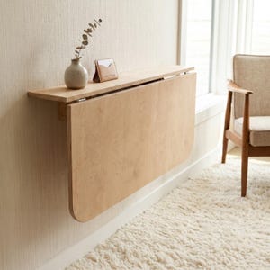 May include: A wall-mounted light wood drop-leaf table with a smooth surface and rounded edges. On the top shelf sits a small, round, light-colored ceramic vase containing a single dried branch and a small, tan, rectangular card holder.