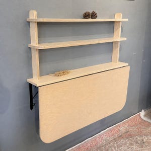 May include: A wall-mounted light wood desk featuring a fold-down surface and two integrated upper shelves. The unit is constructed from plywood with visible grain, supported by black metal brackets, and includes two small pinecones on the top shelf.