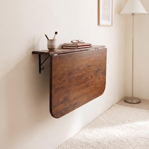 May include: A wall-mounted, dark brown wooden drop-leaf table featuring a rectangular top with rounded corners. The table is attached to the wall with black metal brackets and is shown in a folded-down position. A small ceramic cup with brushes and a notebook with glasses sit on the top surface.
