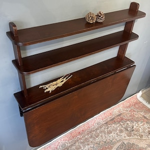 May include: A dark brown wooden wall-mounted unit featuring two upper shelves and a large drop-leaf table surface. The top shelf holds two small pinecones, while the middle shelf displays a small bundle of dried wheat stalks for decoration.