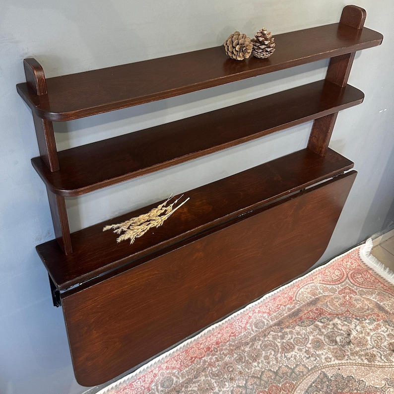 May include: A dark brown wooden wall-mounted unit featuring two upper shelves and a large drop-leaf table surface. The top shelf holds two small pinecones, while the middle shelf displays a small bundle of dried wheat stalks for decoration.