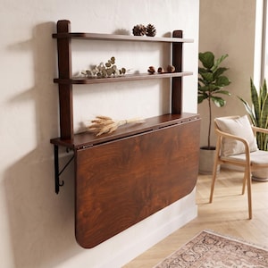 May include: A wall-mounted dark brown wooden unit featuring two upper shelves and a large, hinged drop-leaf table surface. The unit is supported by black metal brackets and displays a smooth, polished wood grain finish throughout the structure.
