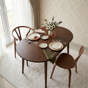 May include: A dark brown wooden oval dining table with two chairs, featuring a centrepiece of white anemone flowers in a vase, two place settings with ceramic plates, silverware, wine glasses, and a folded olive green cloth napkin.