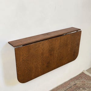 May include: A wall-mounted drop-leaf table featuring a rich, dark brown wood grain finish. The rectangular top is attached to a horizontal support board, with a large, rounded-edge panel that folds downward against the wall surface.