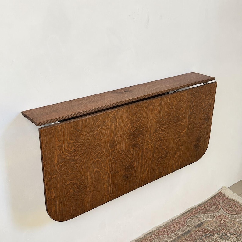 May include: A wall-mounted drop-leaf table featuring a rich, dark brown wood grain finish. The rectangular top is attached to a horizontal support board, with a large, rounded-edge panel that folds downward against the wall surface.
