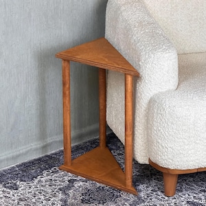 Op de afbeelding: Een driehoekige houten bijzettafel met een warme bruine afwerking. De tafel heeft een driepootontwerp, met een driehoekig boven- en onderblad, en staat naast een witte, getextureerde fauteuil. De hoogte van de tafel is ongeveer 51 cm.