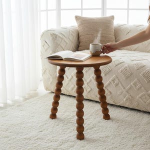 May include: A round wooden side table with three legs composed of stacked spheres. The table surface holds an open book and a two-toned ceramic mug with a light-colored top and a brown base. The table is constructed from smooth, light-brown wood.