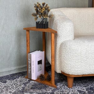 Puede incluir: Una mesa auxiliar triangular de madera con un acabado marrón cálido. La mesa tiene un estante inferior con dos libros y dos piñas. Una pequeña planta en maceta se encuentra en la superficie superior. La mesa está junto a un sofá blanco texturizado.