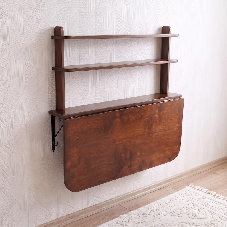 May include: A wall-mounted dark brown wooden unit featuring two upper shelves and a large, rectangular drop-leaf table surface that is currently folded down in a vertical position against the wall, supported by metal brackets on the sides.