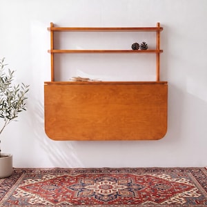 May include: A wall-mounted wooden desk featuring a fold-down rectangular surface with rounded corners and two open shelves above. The unit is crafted from medium-toned wood with a visible grain, providing a functional workspace and storage solution.