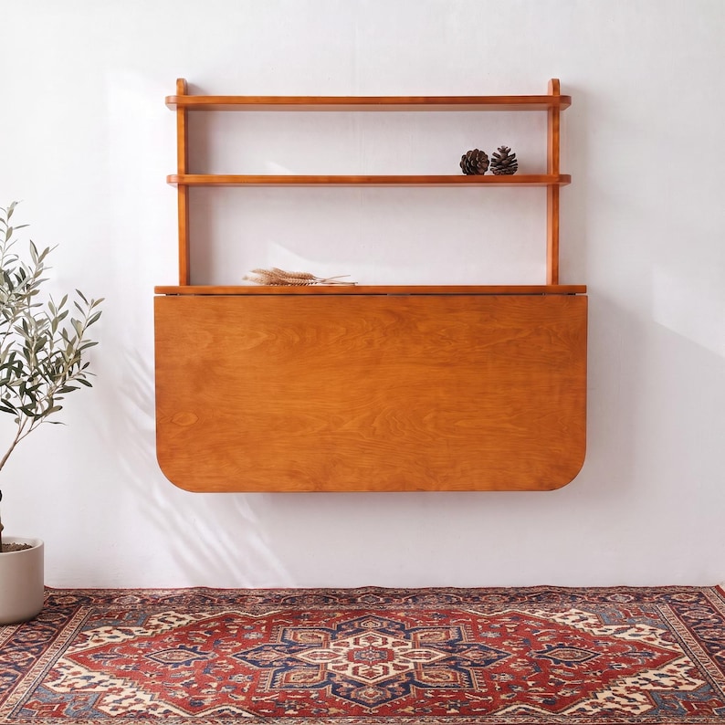 May include: A wall-mounted wooden desk featuring a fold-down rectangular surface with rounded corners and two open shelves above. The unit is crafted from medium-toned wood with a visible grain, providing a functional workspace and storage solution.