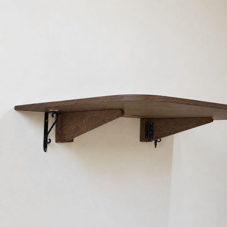 May include: A wall-mounted folding shelf featuring a dark brown wooden surface with a smooth, rounded edge. The shelf is supported by two triangular wooden brackets attached to the wall with black metal hinges and decorative black metal hardware.