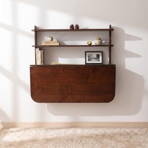 May include: A dark brown wall-mounted drop-leaf desk featuring two open shelves above the folding surface. The shelves hold small decorative items, books, and a framed photograph. The desk surface is smooth with a visible wood grain pattern.