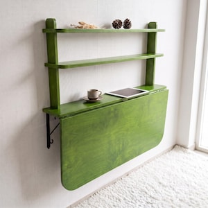 May include: A wall-mounted green wooden desk featuring a drop-leaf table surface and two upper shelves. The desk is supported by black metal brackets and holds a small ceramic cup on a saucer and a tablet computer on its flat surface.