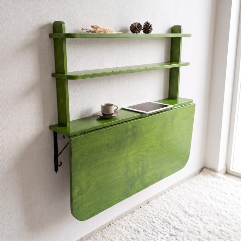 May include: A wall-mounted green wooden desk featuring a drop-leaf table surface and two upper shelves. The desk is supported by black metal brackets and holds a small ceramic cup on a saucer and a tablet computer on its flat surface.