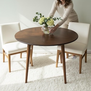 Könnte beinhalten: Ein runder, dunkelbrauner Esstisch aus Holz mit vier abgewinkelten Beinen. Zwei weiß gepolsterte Stühle sind auf beiden Seiten positioniert. Eine klare Glasvase mit Blumen steht auf dem Tisch. Der Tisch hat einen Durchmesser von etwa 107 cm.