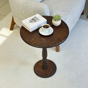 May include: A dark brown, round side table with a pedestal base. A small white teacup and saucer sit on the table, along with a stack of books and a small potted succulent. The table is on a white rug.